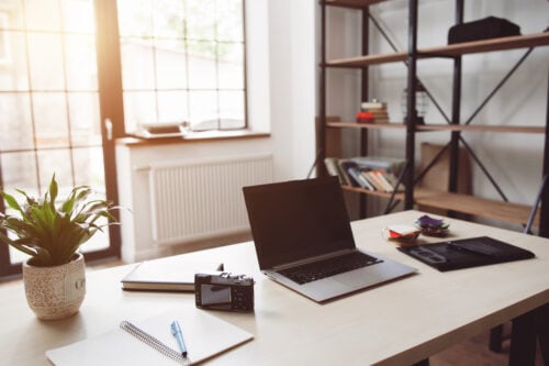 A modern 2023 home office boasts a wooden desk with a laptop, notebook, pen, camera, and tablet. A potted plant adds life on the desk. Shelves filled with books and decor serve as the backdrop, while expansive windows invite in natural light.
