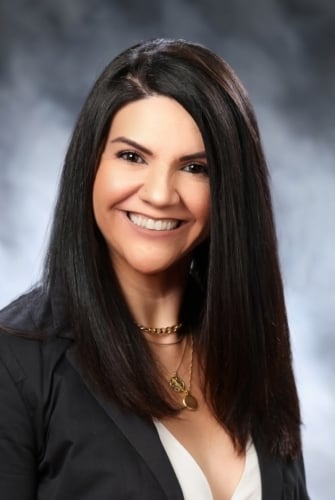 Woman with long, straight dark hair and a bright smile, wearing a black blazer and a white top. She has a necklace, and the background is blurred with shades of grey.