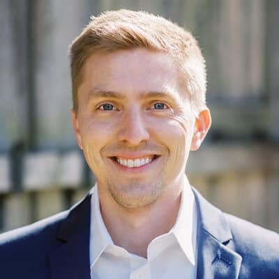 A smiling person with short, light brown hair wearing a dark suit jacket and a white shirt. The background is slightly blurred, showing a wooden fence.