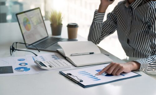 A person in a striped shirt reviews a graph on a clipboard at a desk, pondering the difference between a freelancer and a Virtual Assistant. Nearby, a laptop displays charts alongside an open notebook, calculator, eyeglasses, potted plant, and coffee cup as sunlight filters through the window.