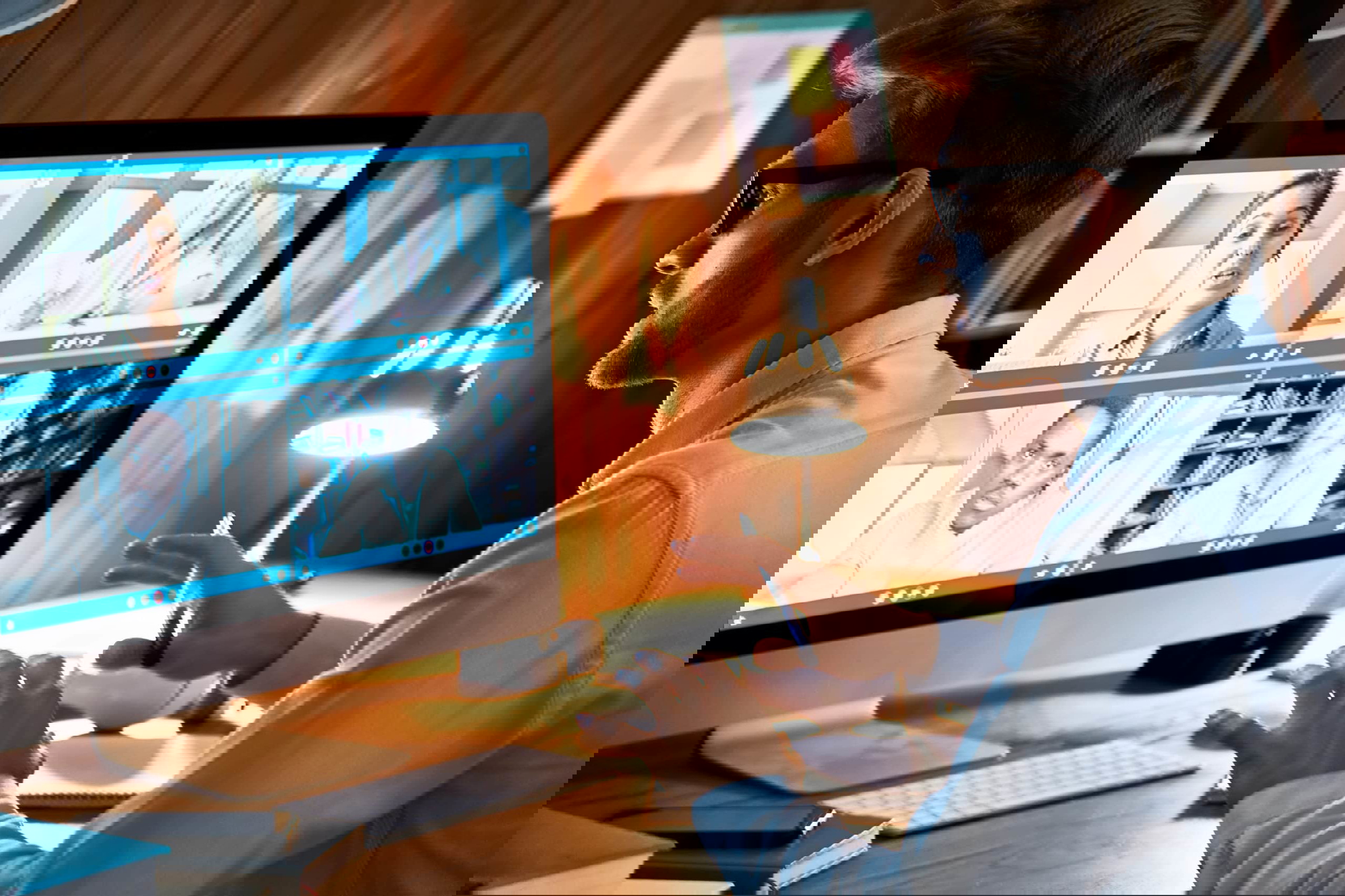 A man in glasses sits at a desk, engaged in a video conference on effective strategies. Four participants are visible, each in their own video window, discussing topics like How To Train Your Virtual Real Estate ISA. The room is lit by a desk lamp, with notes on a bulletin board behind him.