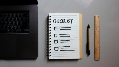 A laptop sits on the left, and next to it is an open spiral notebook titled CHECKLIST with five blank boxes below. A black pen, wooden ruler, and a list of interview questions for hiring virtual assistants complete the scene on the gray surface.