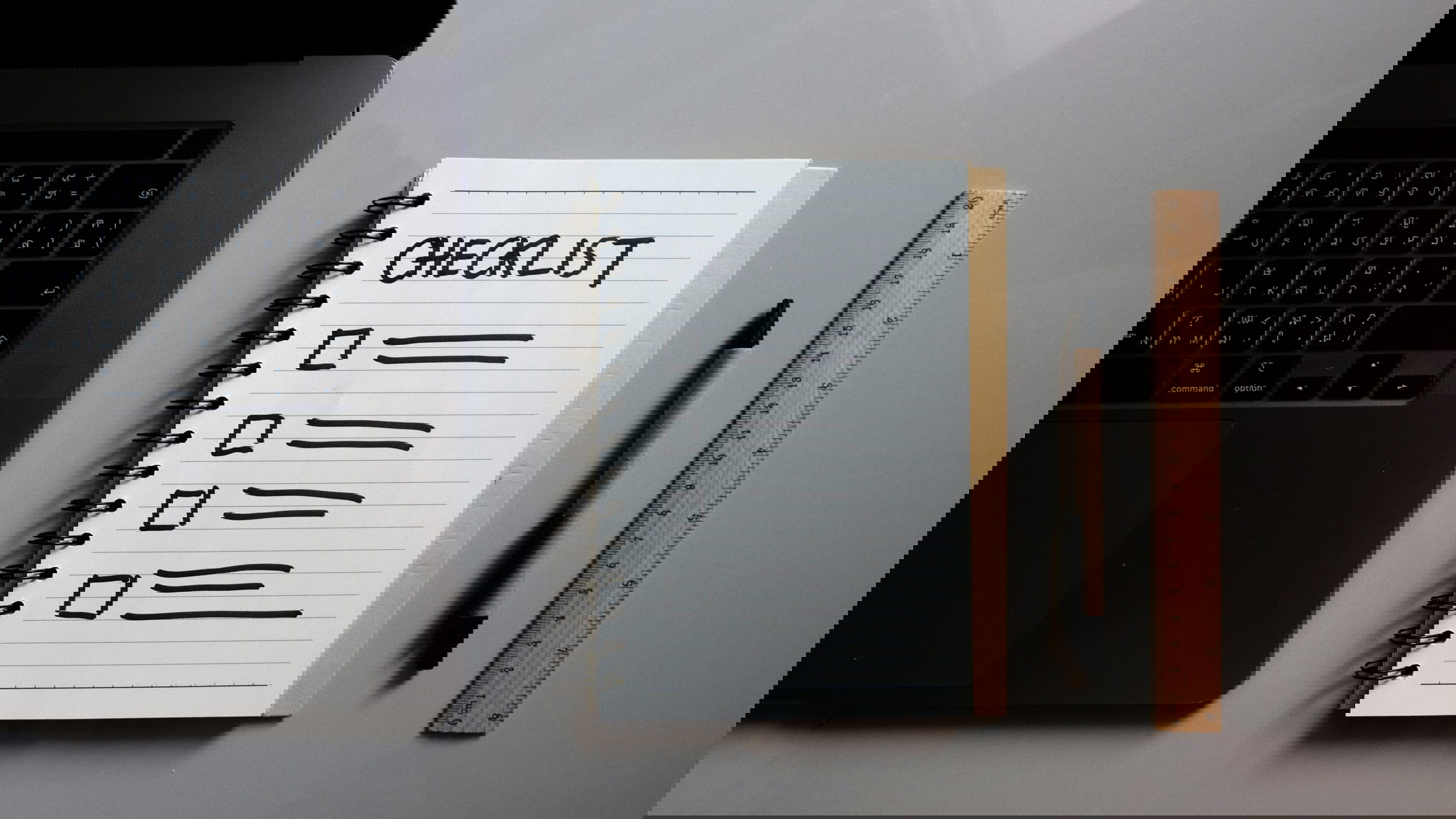 A laptop sits on the left, and next to it is an open spiral notebook titled CHECKLIST with five blank boxes below. A black pen, wooden ruler, and a list of interview questions for hiring virtual assistants complete the scene on the gray surface.
