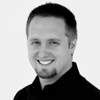 A man with short hair and a goatee, wearing a dark shirt, smiles at the camera in this black-and-white image. His confident demeanor hints at someone like Cliff Stoltzfoos, whose presence commands attention even without uttering a single word.