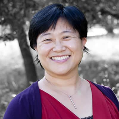 Doreen Tando, with her short dark hair, beams in a red top and dark cardigan, standing outdoors framed by lush trees.