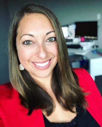 Hallie Warner, a smiling woman with long brown hair, dons a red blazer as she stands indoors against a blurred office background.