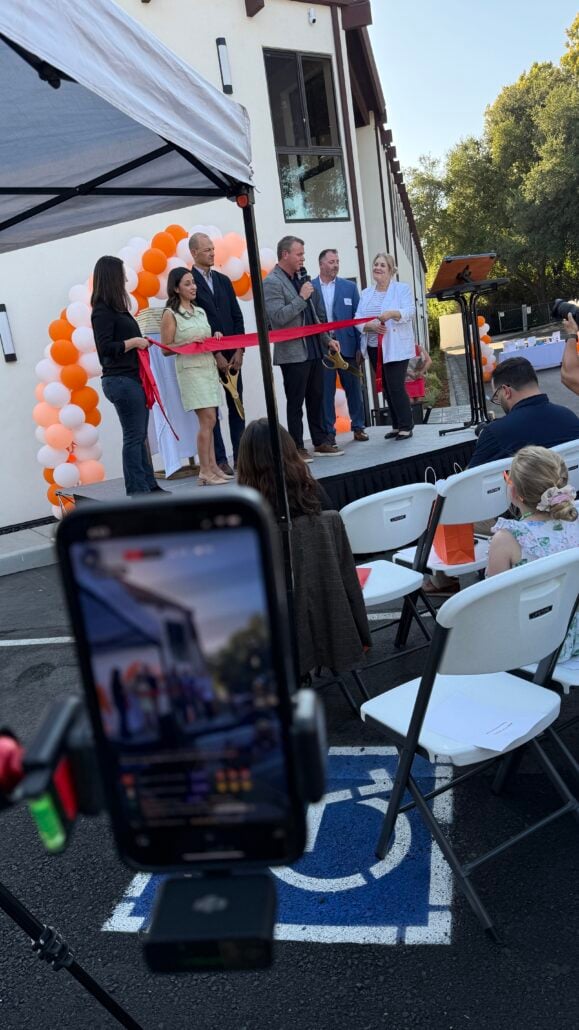 A group of people stand under a canopy at the MyOutDesk Sacramento headquarters grand opening ribbon-cutting event. Orange and white balloons decorate the area. A smartphone on a tripod records the ceremony, while white chairs await attendees.