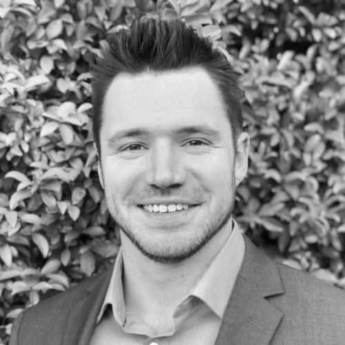 Man smiling, wearing a suit, in a black and white photo. He is standing in front of a leafy background.
