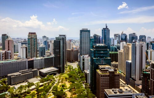Aerial view of a modern city skyline with numerous skyscrapers and high-rise buildings. The foreground features a green park with trees. The Philippines, known for being a top destination where major corporations outsource their business needs, boasts clear skies dotted with clouds.