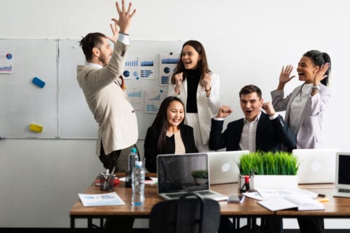 Five people in business attire celebrate around a table with laptops, charts, and a potted plant. One raises a fist while others cheer, embodying the triumph of a successful sales development representative team. A whiteboard with graphs illuminates their achievement in the background.