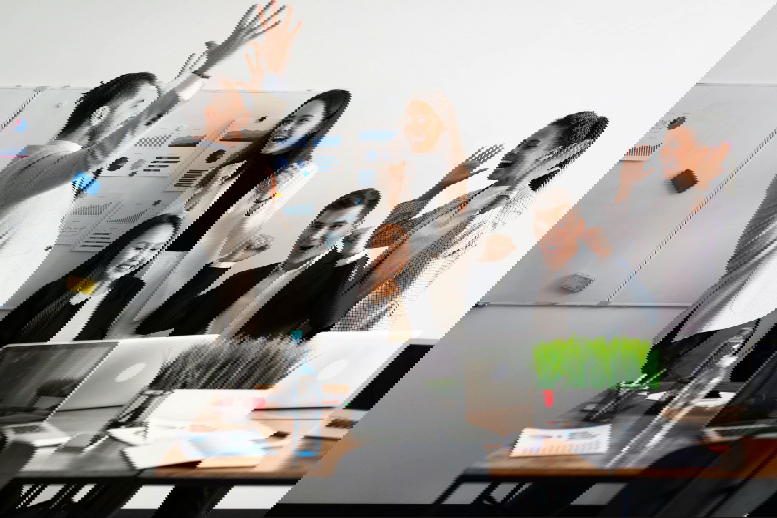 Five people in business attire celebrate around a table with laptops, charts, and a potted plant. One raises a fist while others cheer, embodying the triumph of a successful sales development representative team. A whiteboard with graphs illuminates their achievement in the background.