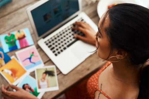 A woman in a red polka dot dress works on a laptop at a wooden table, exploring Fancy Hands alternatives. Colorful photos are spread out beside her. She has dark hair tied back and is wearing hoop earrings.