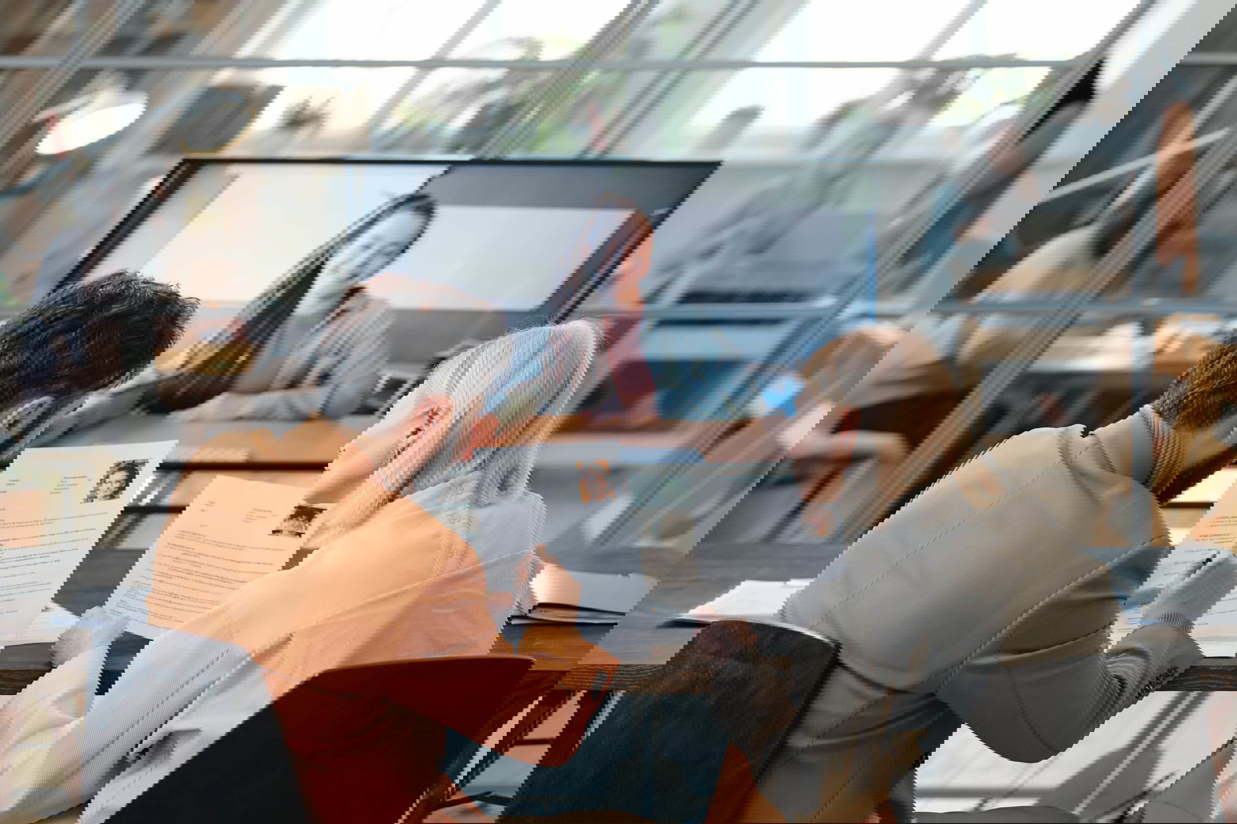 Two people sit at a table with resumes, conducting a video interview in the bright, modern office. The candidate appears on a large screen in this guide on how to hire virtual employees and why you should. Through the glass wall, more colleagues are visible working diligently in the background.