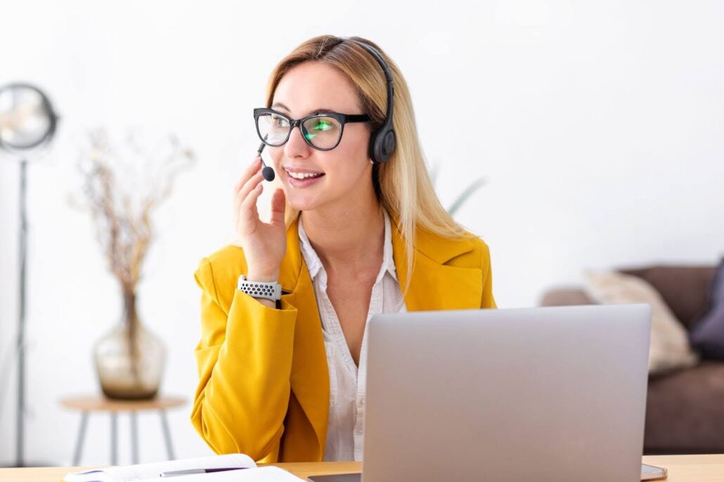 A woman with blonde hair and glasses, wearing a yellow jacket, is using a headset at her desk with a laptop. She embodies the essence of a virtual receptionist, known for engaging interactions. The blurred background features a floor lamp and decorative plant as she smiles warmly.