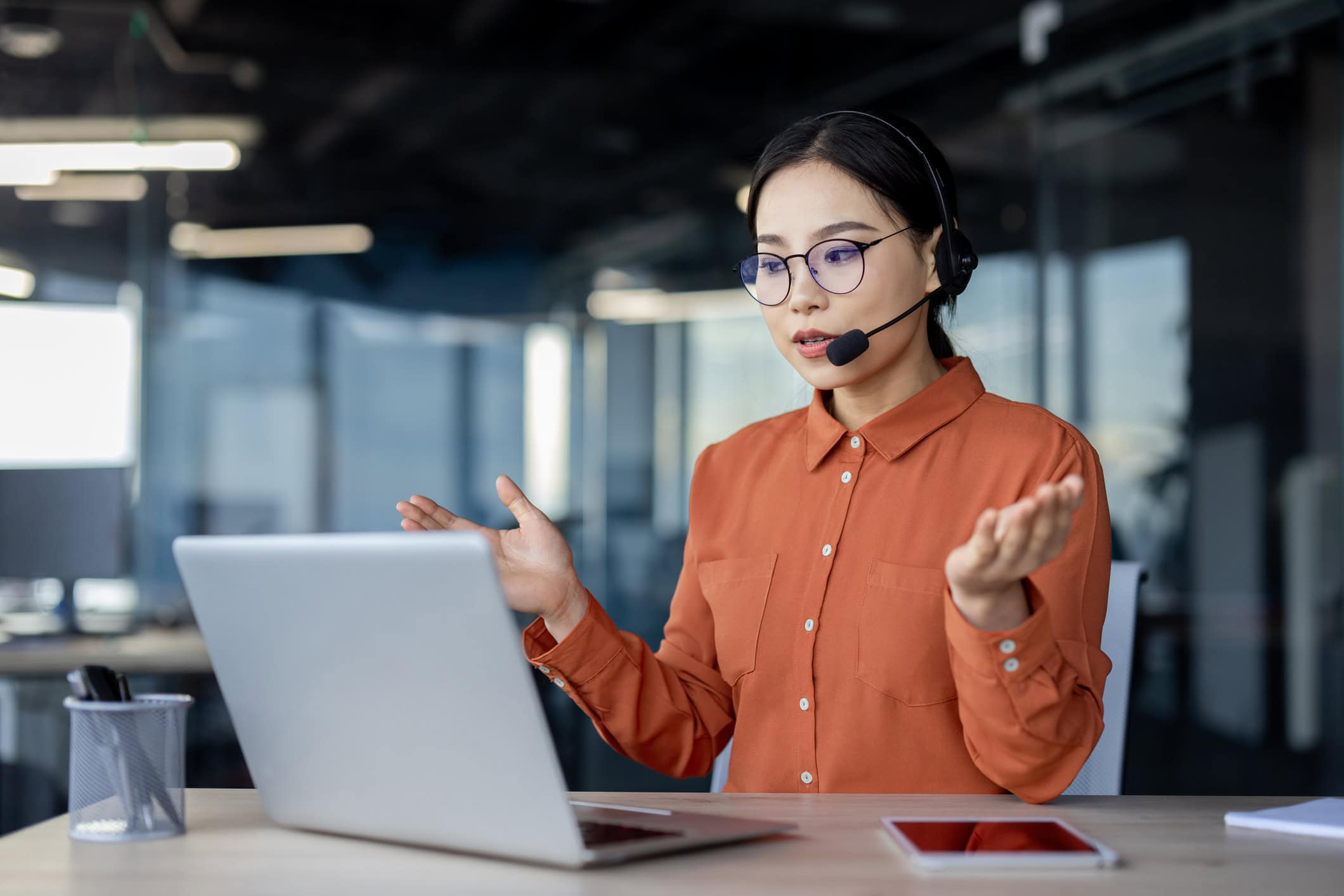 Friendly businesswoman wearing a headset, smiling while assisting customers via video call on her laptop, working as a help desk professional in an office setting.