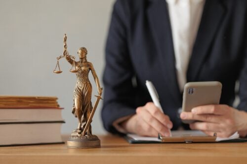 A statue of Lady Justice with balance scale and sword takes center stage on a desk. Behind her, a person in a suit is holding a smartphone and writing on a clipboard, perhaps noting the benefits and opportunities law firms gain when hiring virtual legal assistants. Nearby are stacked books.