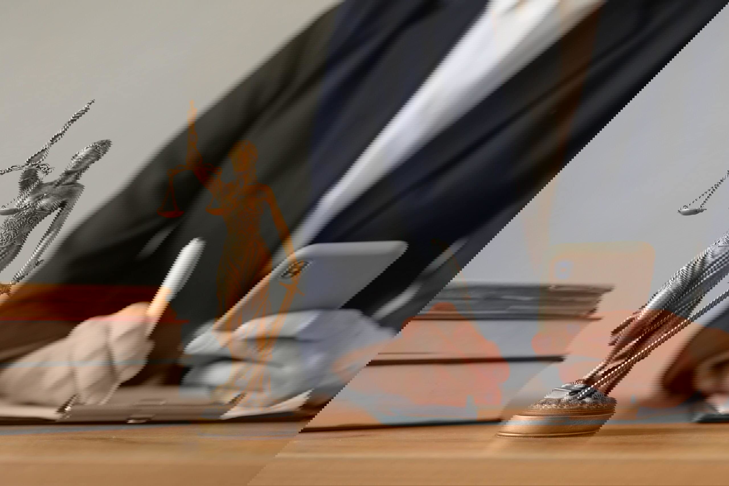 A statue of Lady Justice with balance scale and sword takes center stage on a desk. Behind her, a person in a suit is holding a smartphone and writing on a clipboard, perhaps noting the benefits and opportunities law firms gain when hiring virtual legal assistants. Nearby are stacked books.