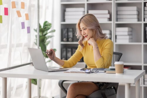 A person with blonde hair in a yellow sweater sits at a desk, engrossed in researching Zirtual alternatives on their laptop. The desk is cluttered with papers, glasses, a pen, and a coffee cup. Shelves filled with boxes and binders line the background, while colorful sticky notes adorn the window.