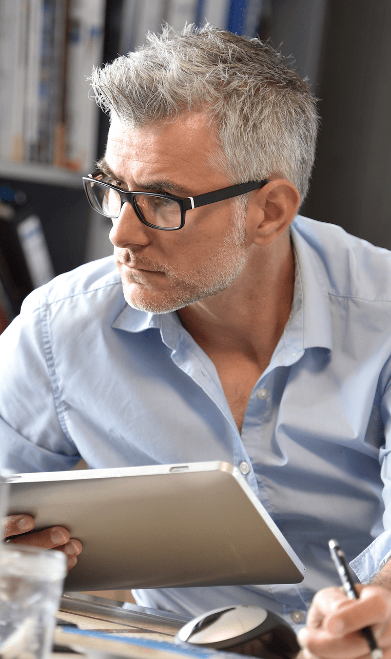 A man with gray hair and glasses is attentively looking to the side while holding a tablet, showcasing MyOutDesks enterprise solutions. Dressed in a light blue shirt, he sits at a desk, pen in hand, surrounded by various office items.