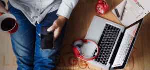 Person holding coffee and smartphone seated on floor. Laptop with open book, red headphones, notebooks, and red alarm clock nearby. Wearing blue jeans and checkered shirt. Desk surface and tech equipment depicted from above.