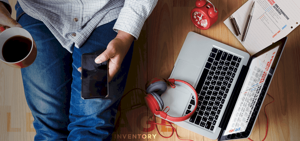 Person holding coffee and smartphone seated on floor. Laptop with open book, red headphones, notebooks, and red alarm clock nearby. Wearing blue jeans and checkered shirt. Desk surface and tech equipment depicted from above.
