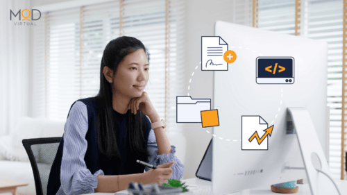 A woman sits at a desk with a computer, holding a pen. Glancing at icons of documents, a folder, and a coding window on her screen, she ponders the question: What is a Healthcare Virtual Assistant? The room is bright with white shutters.